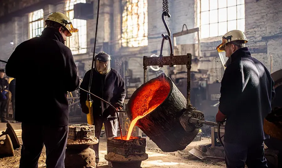 Three people stand inside a factory casting a large bell. Molten metal pours from a container held in the air by a hoist