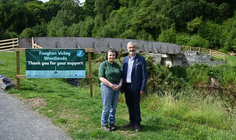 Two people are stood in front of the new bridge in Faughan Valley woodlands. Next to them is a sign to introduce the woodlands and has the Heritage Fund logo on it.