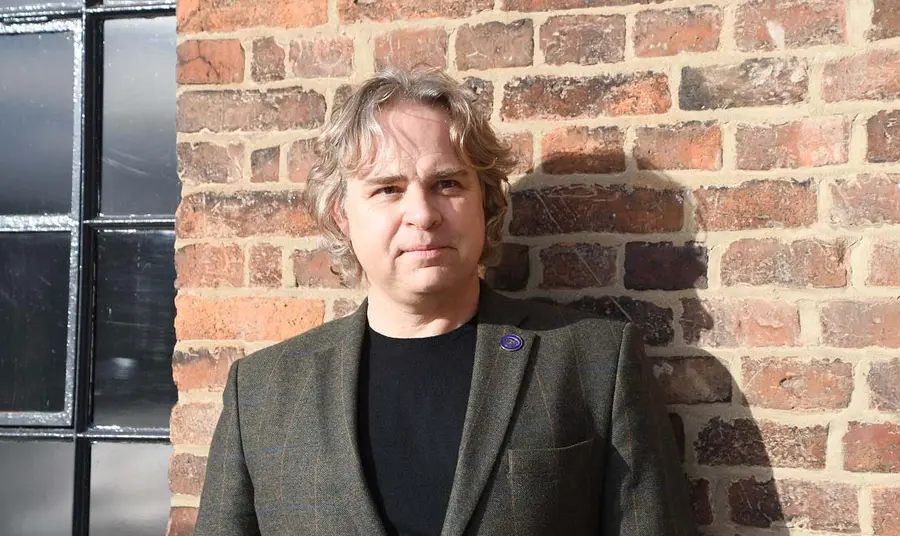 A person wearing a black and grey suit, standing in front of a red brick wall