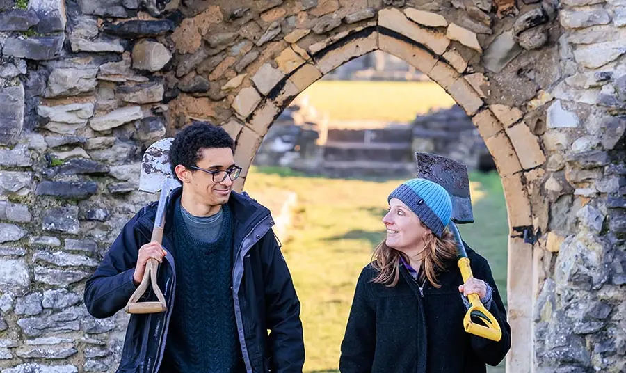 Two people holding shovels and dressed for cold weather walk through a doorway in a wall