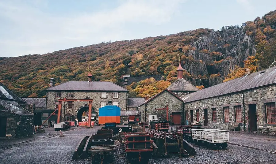 cwrt Amgueddfa Lechi Cymru yn Llanberis, gogledd Cymru, gydag adeiladau ffatri cerrig o amgylch iard gydag offer diwydiannol ar gyfer prosesu llechi, gyda'r mwynglawdd llechi i'w weld yn y cefndir