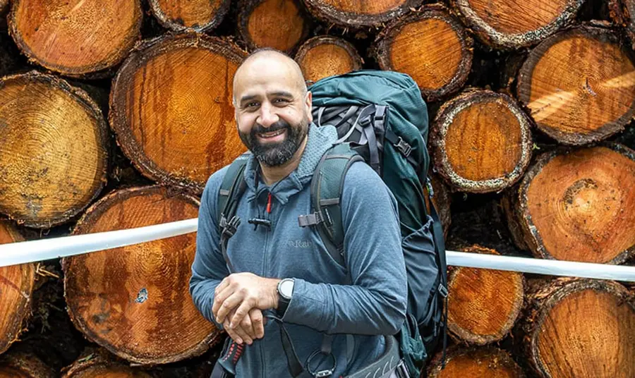 A man from a Pakistani family background on a outdoor walk, wearing hiking gear. He stands in front of a pile of logs