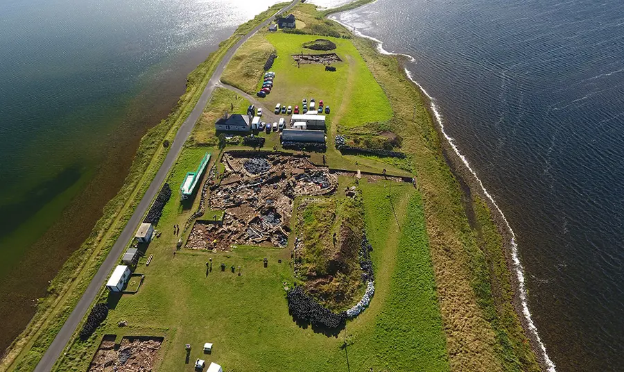 The Ness of Brodgar Looking South East over the site, a finger of grass covered land with water either side and a thin causeway visible at the top of the image. Trench J in bottom left; Trench P in centre, and Trench T in distance