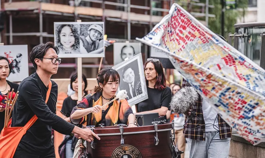 Two people of Chinese heritage play a drum in a procession, while people in the background hold placards with photos of Chinese relatives