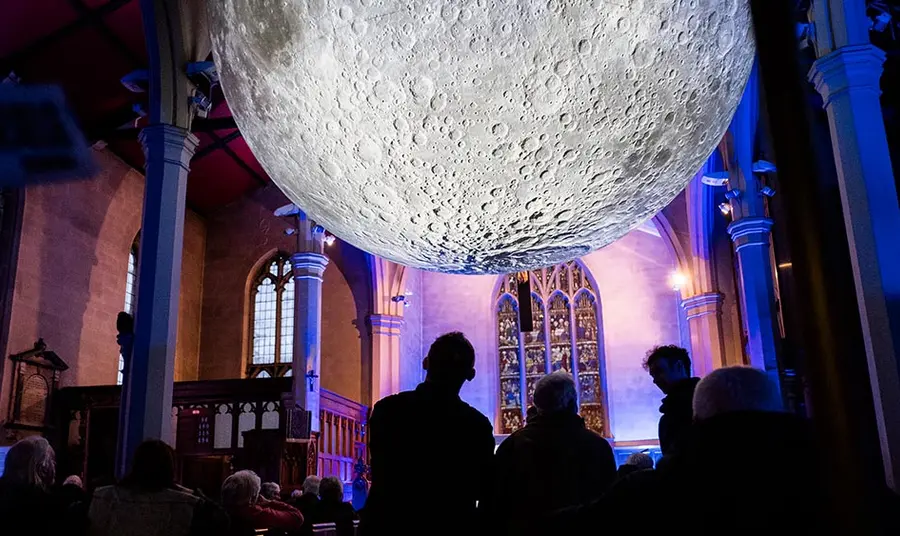 People look at a huge illuminated moon artwork suspended in a medium-size church