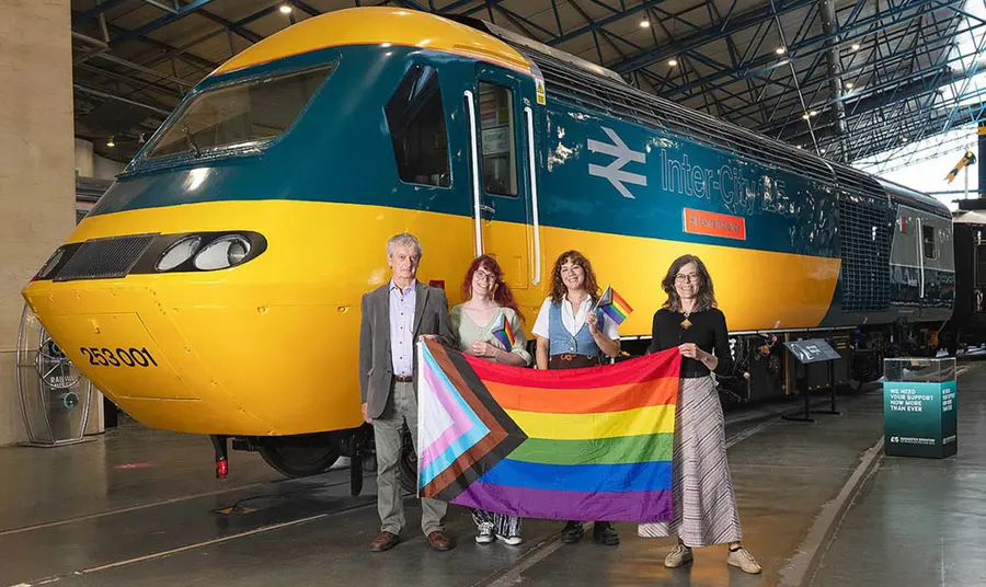 Four people stand in front of a train engine and hold a progress pride flag
