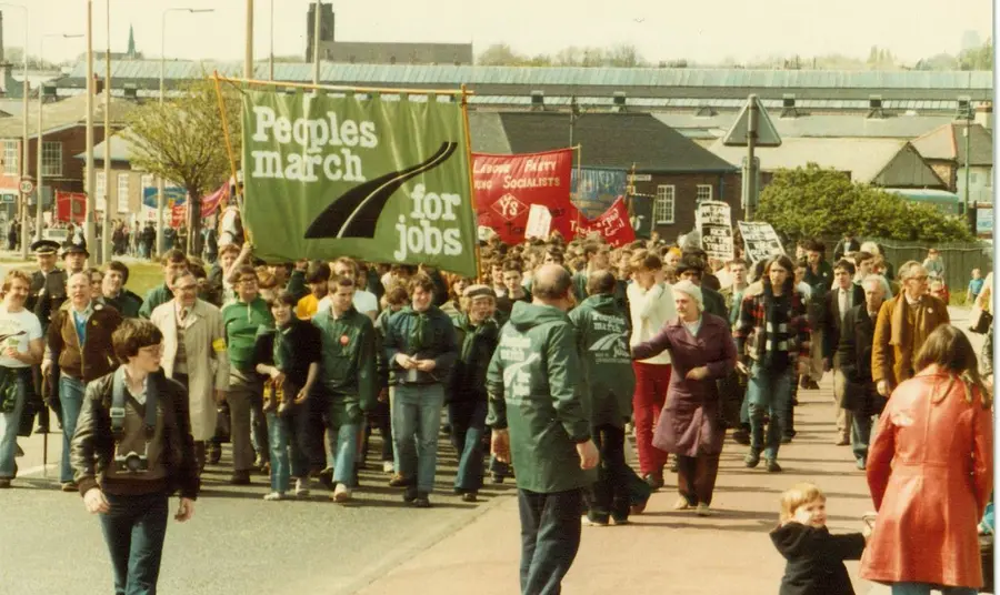 A group of people on a march through an industrial areas. They are holding a large banners, one reads 'People's March for Jobs' 