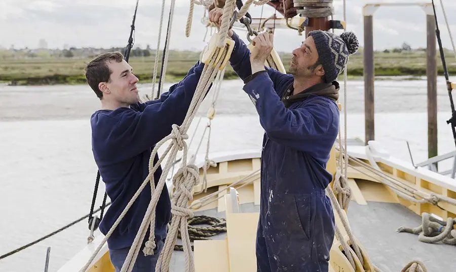 Two people pulling a rope on a historic ship, with a river estuary in the background