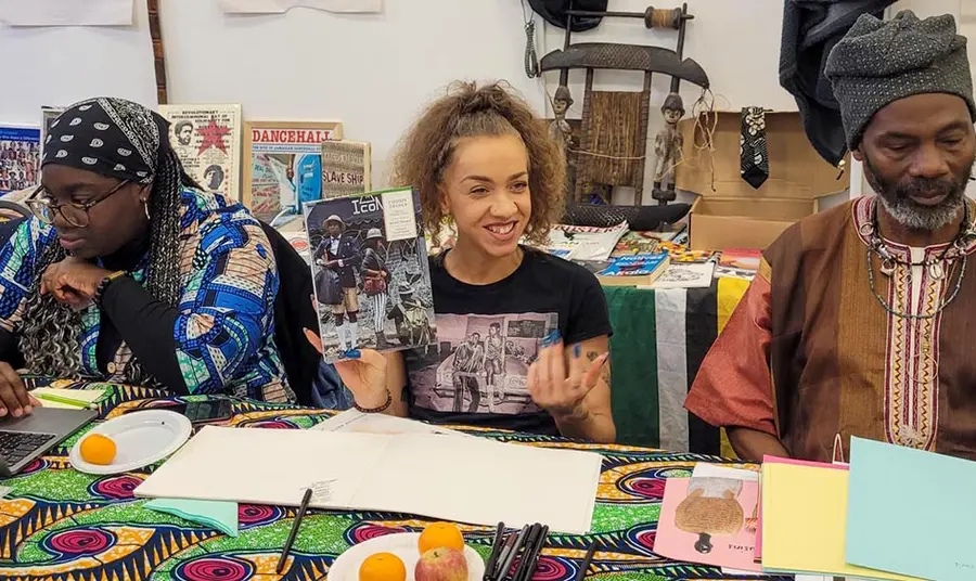Three black people take part in a workshop on black history. All three people have dark skin of various shades, the focus of the image is a woman holding a magazine with black people on the cover