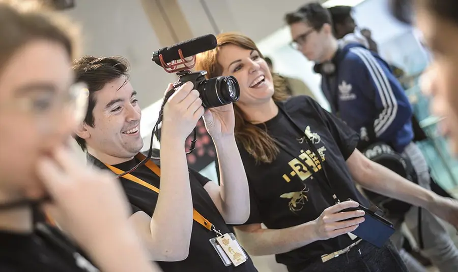 Two people wearing Reimagine, Remake, Replay tshirts using filming equipment at an event