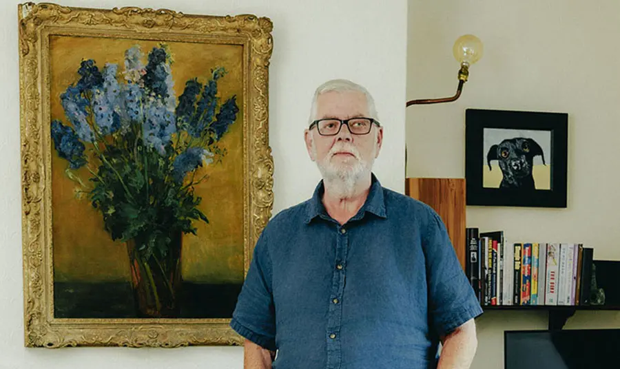 Richard Kennedy stands in a living room in front of paintings and a bookshelf