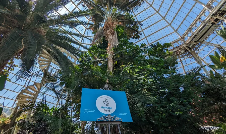 Palm trees and plants inside a glassed roofed building. A board with the Heritage Fund logo is on display on an easel. 