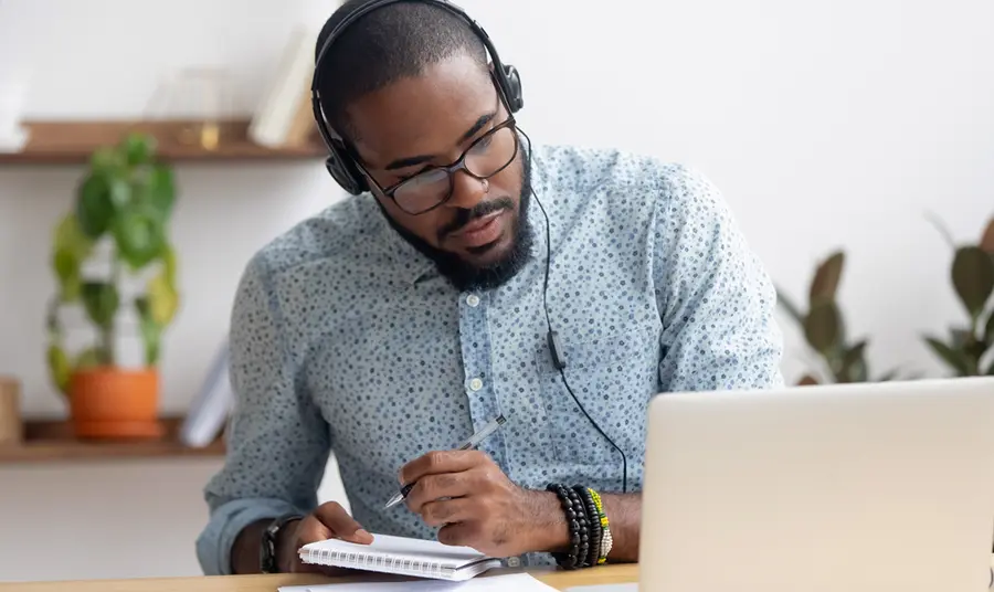 A black man waering headphones, looking at a laptop computer and taking notes in a notebook