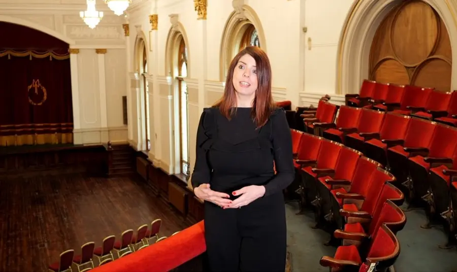 A woman in a black dress standing in an empty theatre speaking to the camera. Red chairs and a curtained stage can be seen in the background.