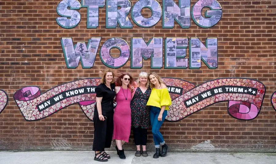 People standing in front of a ceramic mural that spells out the words Strong Women