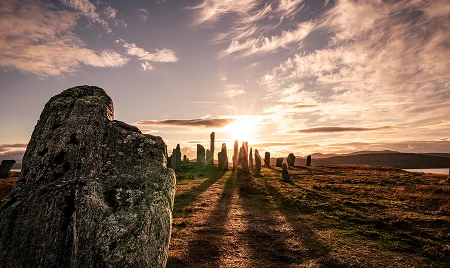 Calanais Standing Stones In the foreground to the left is a standing stone long shadows reach out to it from a group of standing stones behind. The sun is setting with orange tinged light filtering through the cluster of stones