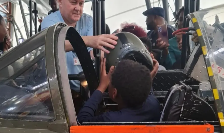 Photograph of a young person sitting in the cockpit of a plane. Someone is passing him a helmet.