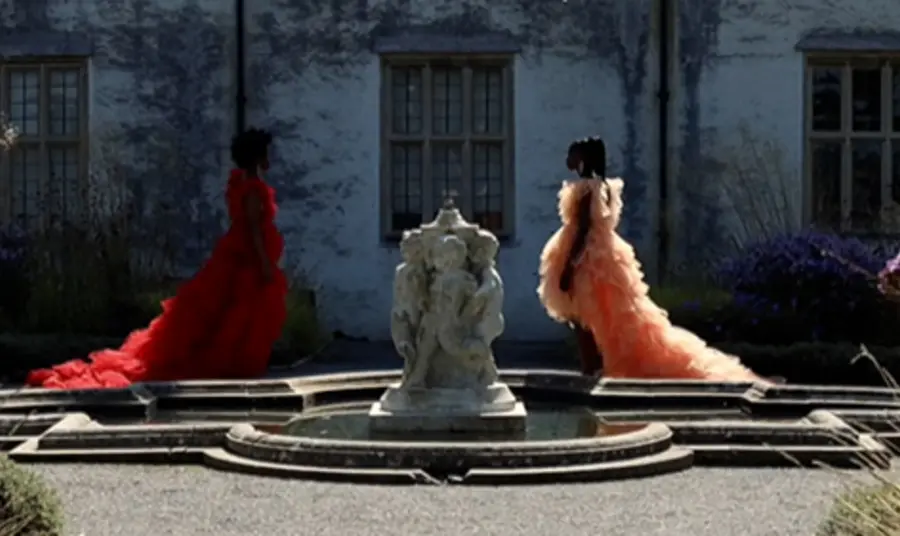 Two people dressed in colourful dresses walk towards a fountain outside a historic house