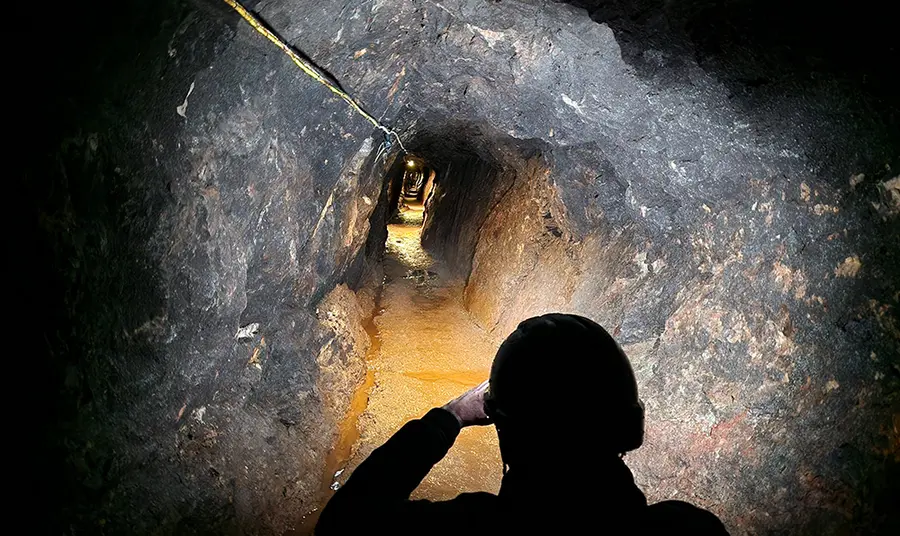 A rock walled mineshaft illuminated by torchlight stretched into the distance, in the foreground the silhouette of a person wearing a miners helmet and looking along the tunnel