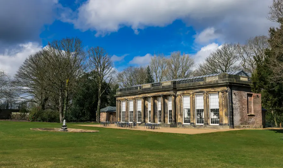 A Georgian stone building with glass roof and large windows. A lawn and stone fountain are in front of the building