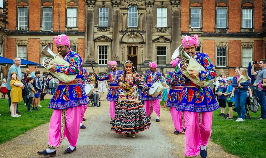 An Asian brass band, with musicians and dancers dressed in colourful clothes, walking down a path in front of a stately home