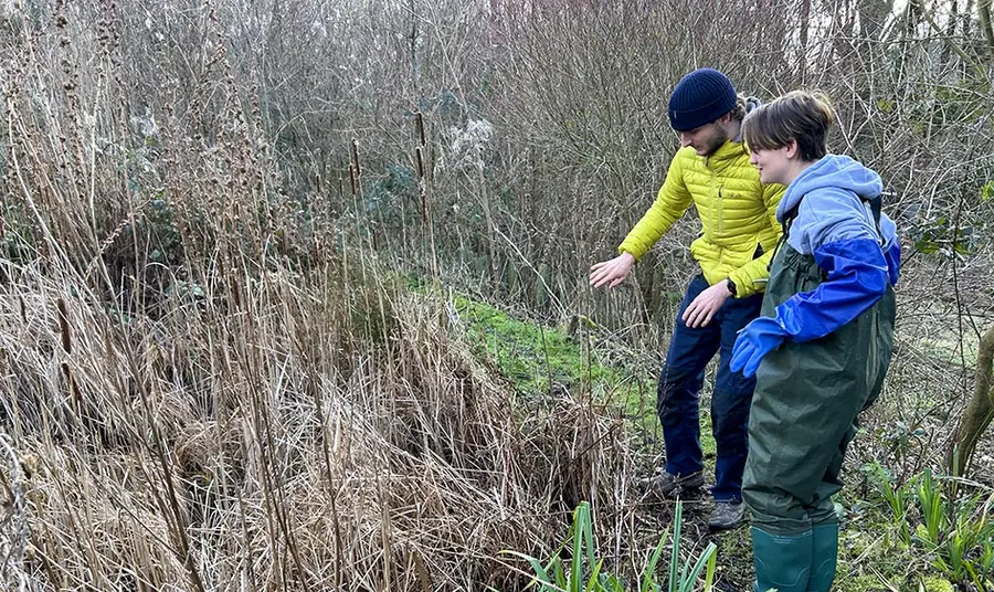 Two people look at an overgrown pond, one person is wearing waders