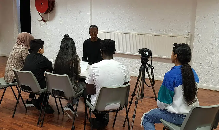 A group of young people sit a table interviewing another young person, with another person to the side operating a camerawith