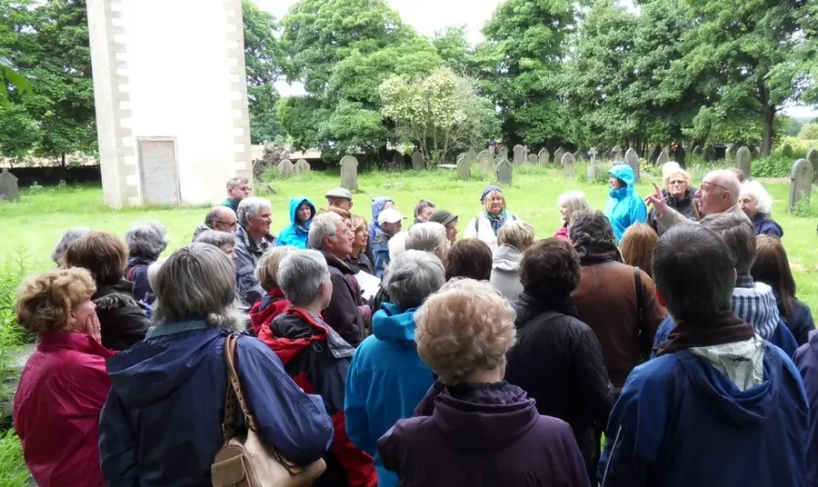 A group of people wearing outdoor clothes take part in a churchyard heritage walk at St Matthew's Churchyard
