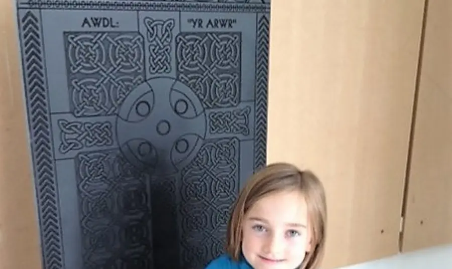 Young child sitting on a high backed wooden chair depicting an intricate carving of a cross. 