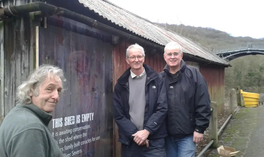 Three people stand outside a large closed shed with a sign saying awaiting restoration
