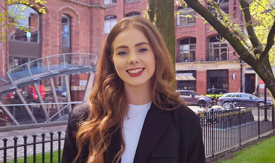 A young white woman with long red-blonde hair wearing a white top and black jacket standing in an urban area.