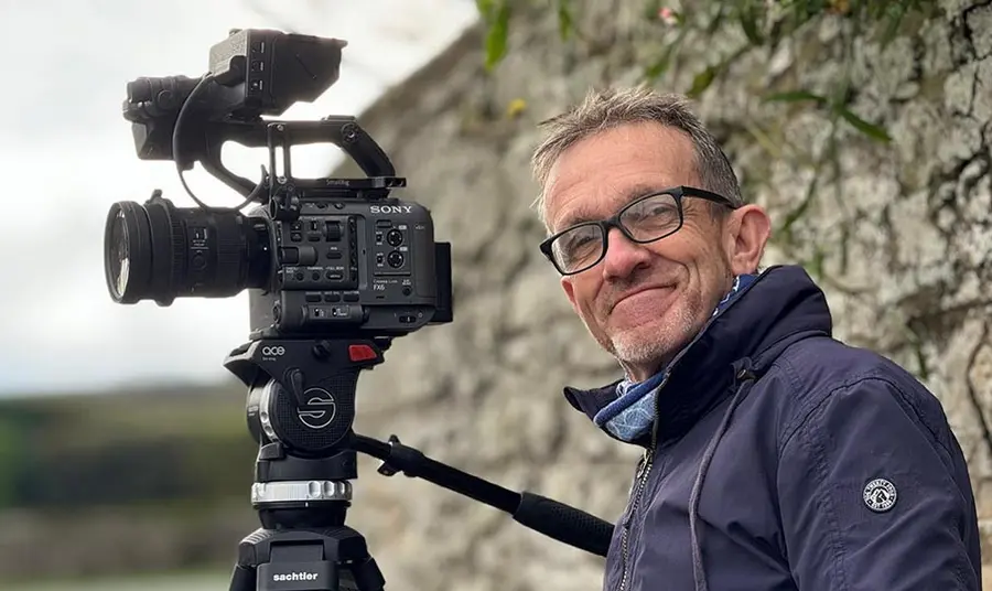 A photograph of a white man standing outside next to a film camera