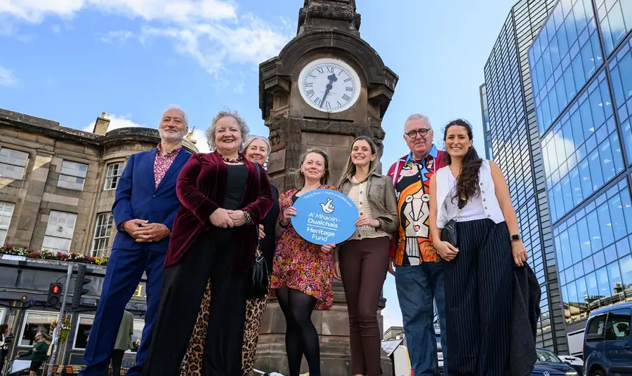 A project group of seven people pose for a photograph in front of the Hear of Midlothian Memorial Clock. Karen Mailley stands at the centre of the group, holding a Heritage Fund branded roundel.