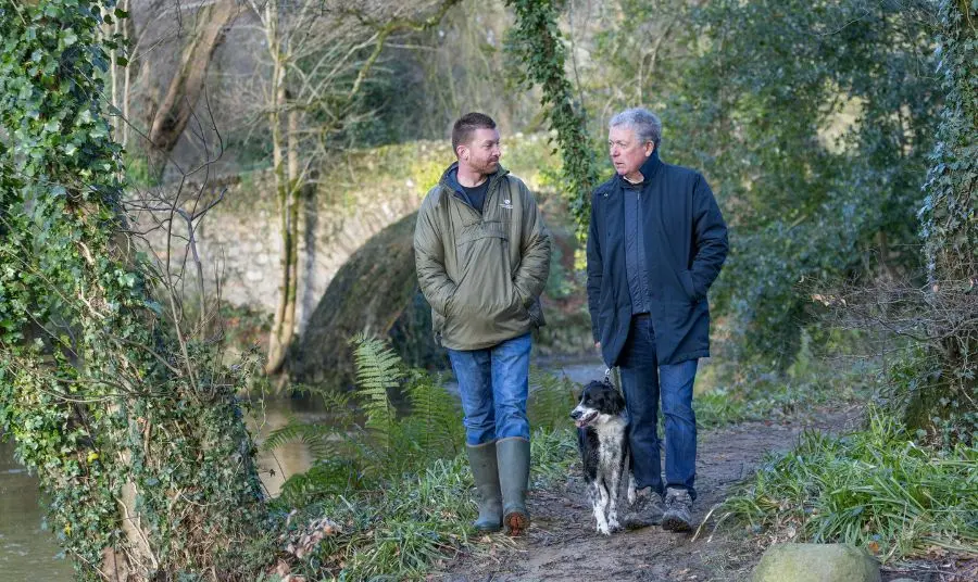 White Water Bridge Mourne Park Two people and a dog are chatting and walking in woodland, behind then is a stone bridge.