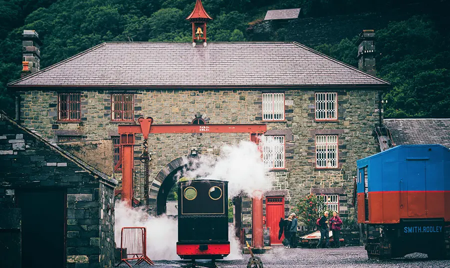 A small train billowing steam in the yard of the museum buildings