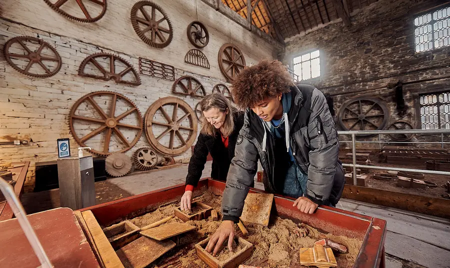Two people do an interactive activity inside an industrial building at the National Slate Museum. In the background large cart wheels hang on the wall.