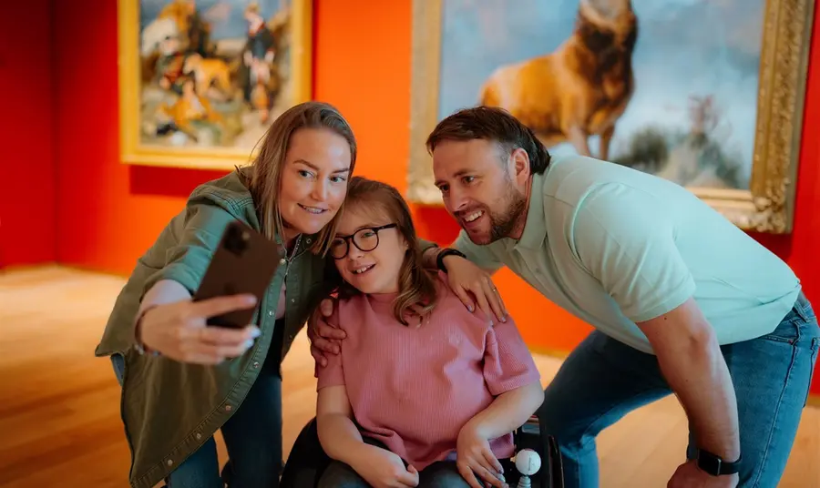 Visitors to the Scottish galleries meet Monarch of the Glen. Three visitors, including a young person in a wheelchair, take a selfie in front of the famous Monarch of the Glen painting.
