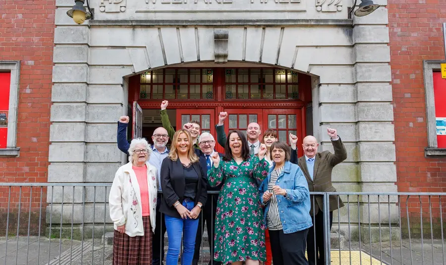 A group of different ages from the Tylorstown community celebrate outside the Welfare Hall, smiling and raising their hands in the air.