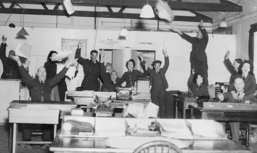 A black and white photograph showing RAF staff celebrating in an office, throwing their arms and papers in the air.