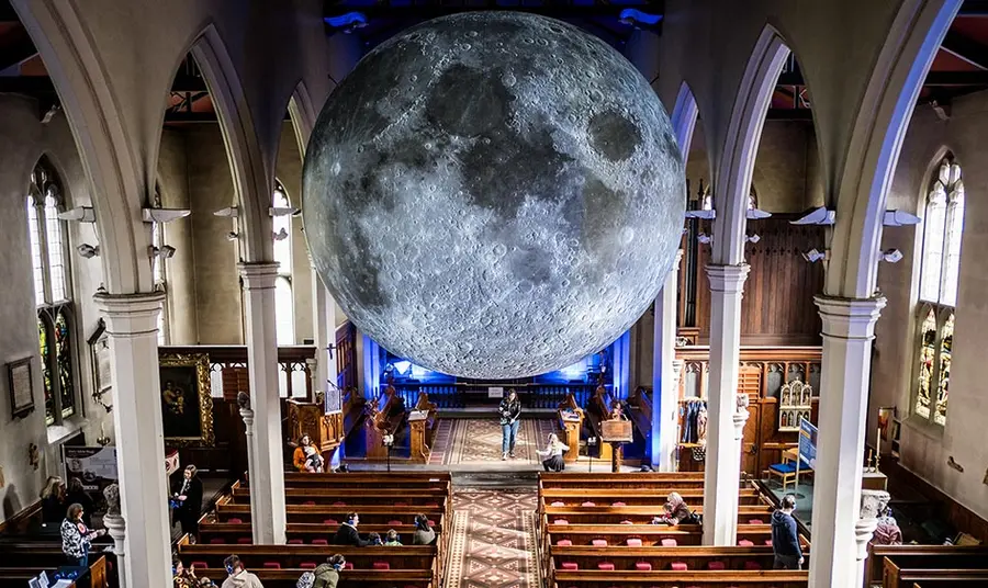 the inside of a medium size English church, with pews and stone columns and an enormous moon suspended from the ceiling
