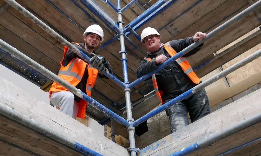 Jordan and an older male colleague, both wearing hi-vis jackets and hard hats, stand above on scaffolding, smiling down.