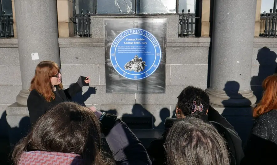 A guide introduces a walking tour group to a blue plaque on the facade of a former community bank in central Airdrie.