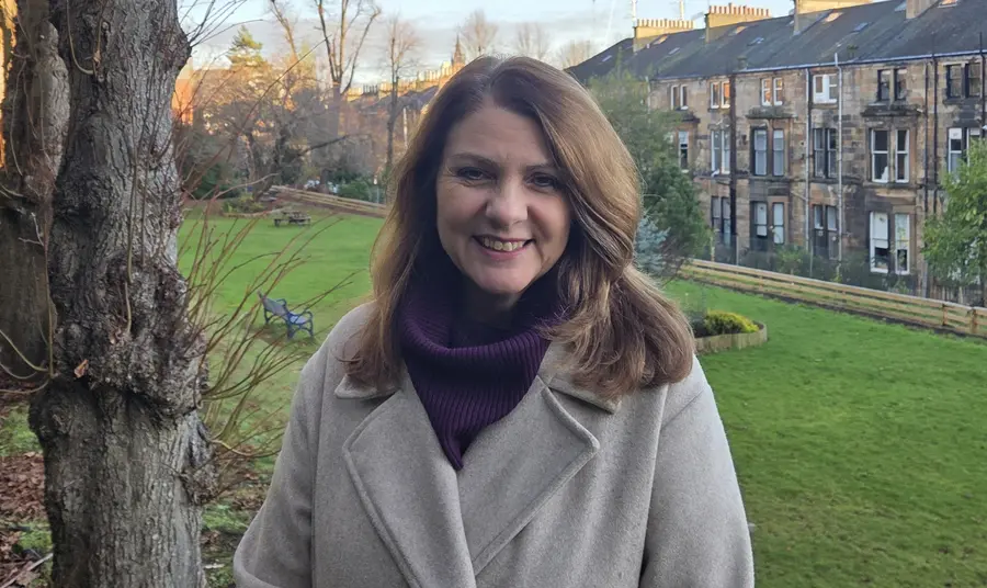 Heather Reid, wearing a grey woollen coat and purple scarf, stands smiling in a field behind a row of Scottish tenement houses.