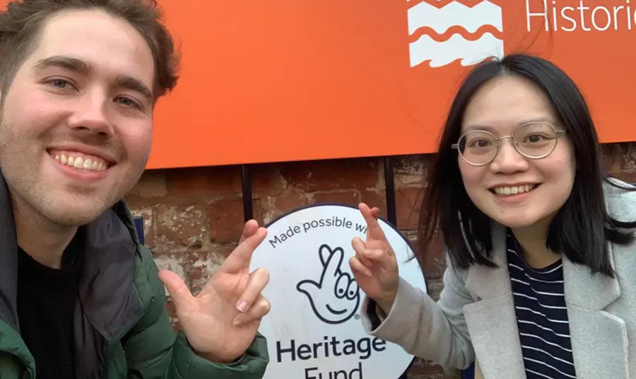 Two people smile and pose with crossed fingers in front of a Heritage Fund acknowledgement plaque