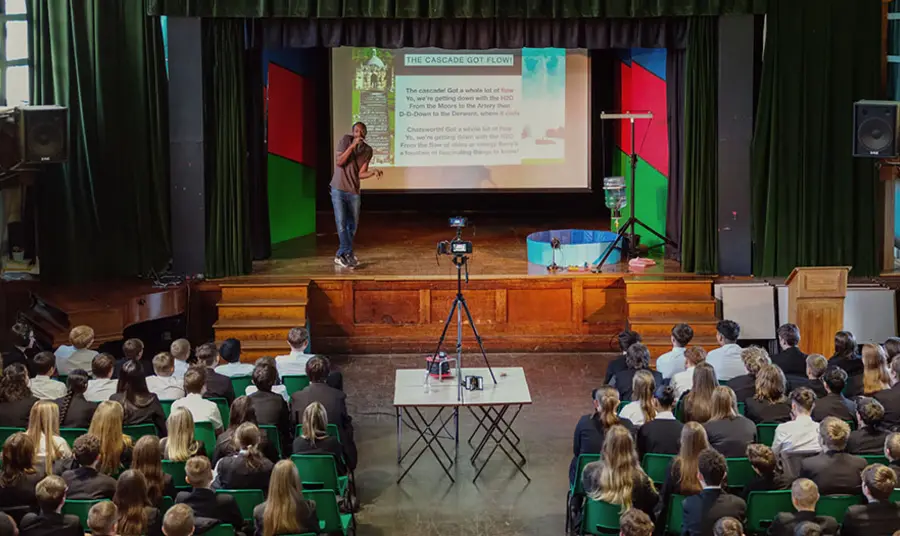 A person is on stage presenting a PowerPoint to a hall full of school children.