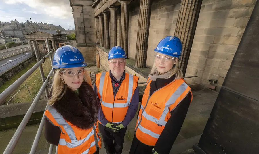 Three people wearing hi-viz jackets and hard hats stand amind the classical columns of the former Royal High School. The City of Edinburgh can be seen behind them