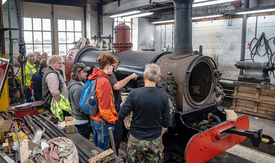 A group of people in a workshop watching someone restoring a steam engine