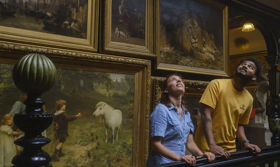 Two people stand on a staircase looking up, with paintings covering the wall behind them