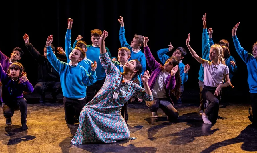 A group of school children on a stage take tuition in Indian dance from an instructor, all striking the same knelt pose together.