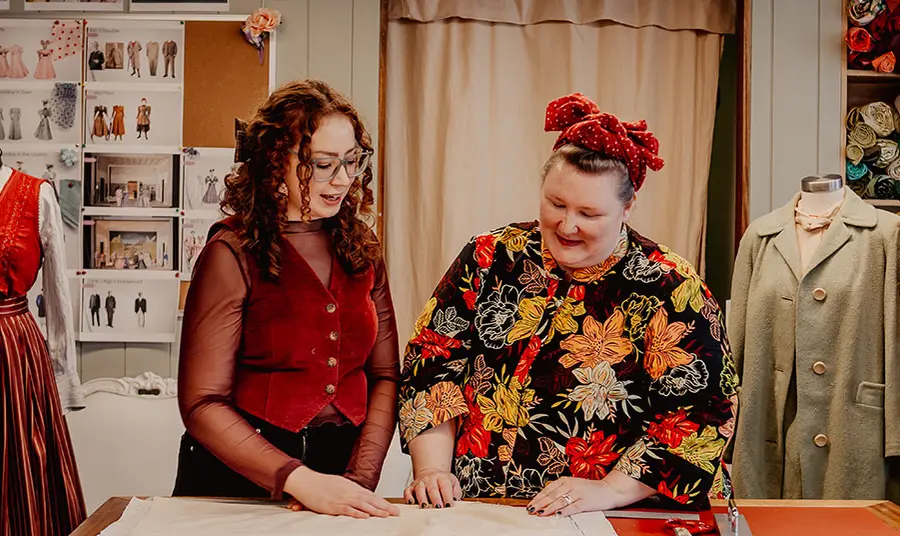 Two people are in a costume room and are looking down at a pattern on a cutting table. In the background are images of costumes, costumes on mannequins and rolls of fabric stacked on shelves
